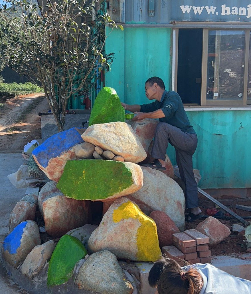 Two people painting the pile of boulders beside the lodge in green, blue and yellow, with the tea rows behind