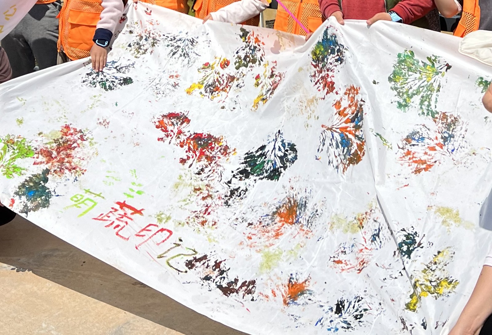 A large white cloth held up by many hands, covered in leaf prints pressed on by a visiting school group