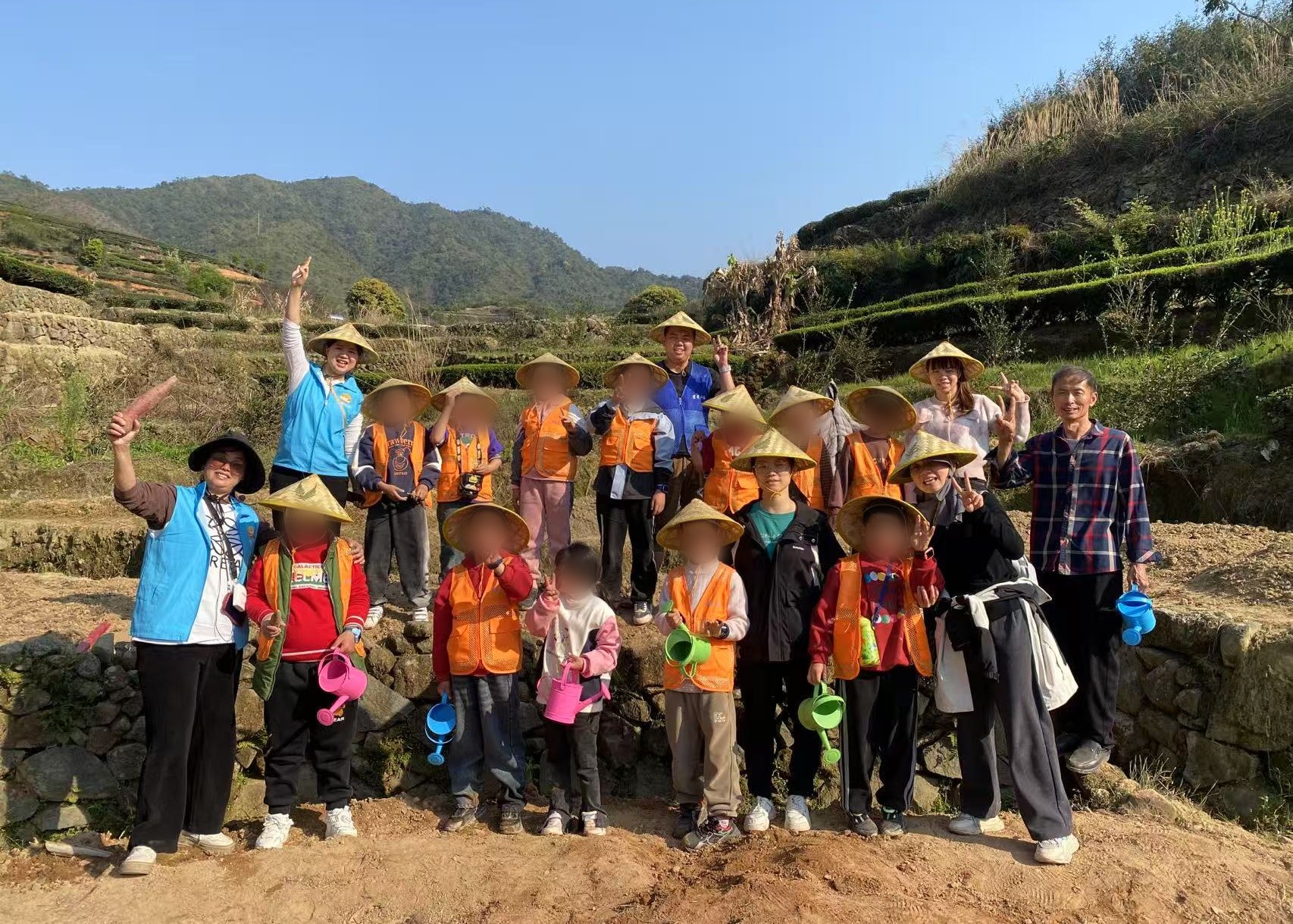 Children in orange vests and bamboo hats standing with staff and volunteers on the tea terraces above the lodge, hills behind them