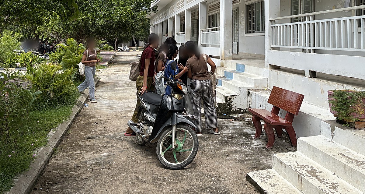 Students standing together beside a motorbike on the path outside the classroom block, faces blurred