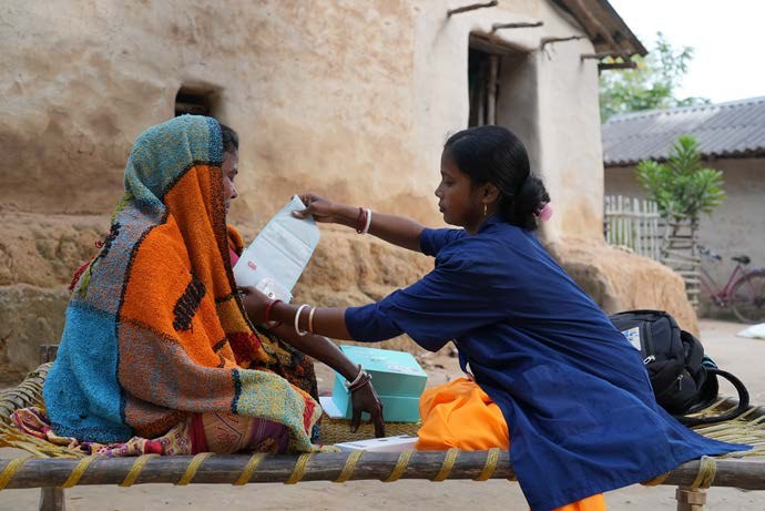 An iKure health worker handing medicine to an elderly woman sitting outside her mud-walled house