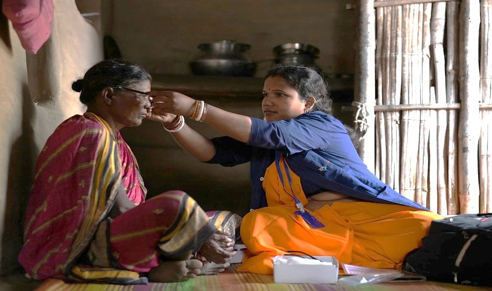 An iKure health worker sitting on the floor of a house, adjusting an elderly woman's spectacles