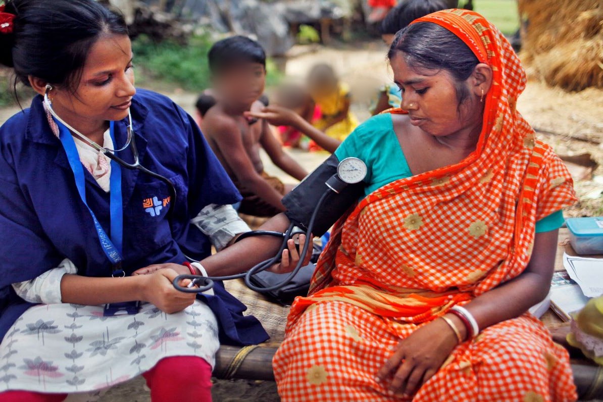 An iKure community health worker taking a woman's blood pressure on a bed outside her house in the village
