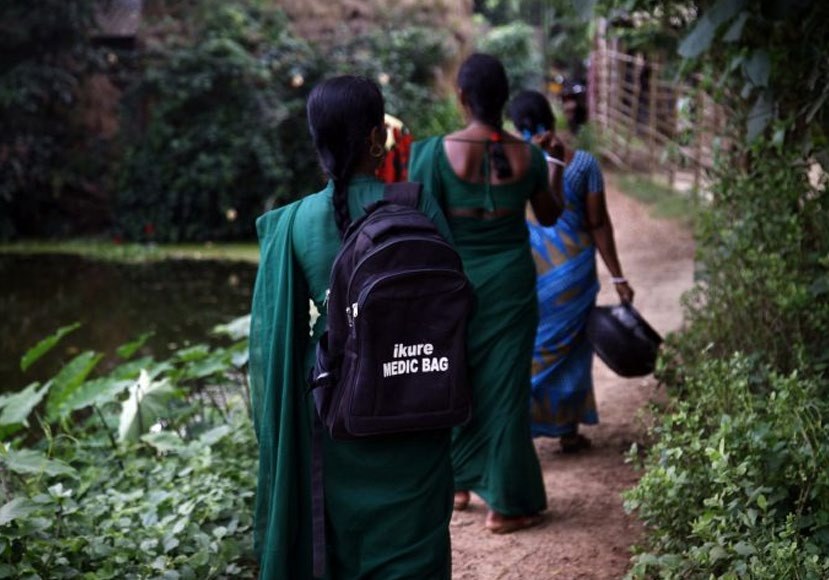 Three iKure health workers walking a village path, the nearest one carrying a backpack marked iKure MEDIC BAG
