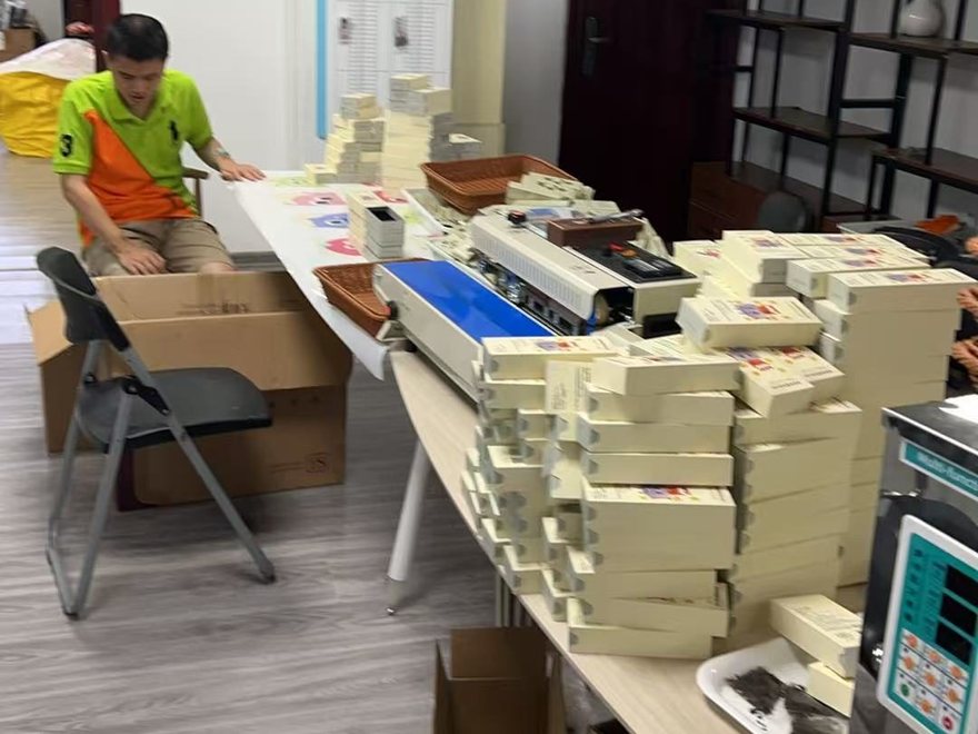A member of the Aihui tea studio at the long packing table, drawing out a roll of printed box wraps, with a sealing machine, scales and stacks of finished gift boxes beside him